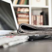 Laptop und mehrere Zeitungen auf einem Tisch, im Hintergrund Bücherregal mit sichtbaren Büchern.