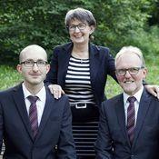 Drei Personen in Anzug und Bluse posieren lächelnd im Freien, umgeben von Grünpflanzen.